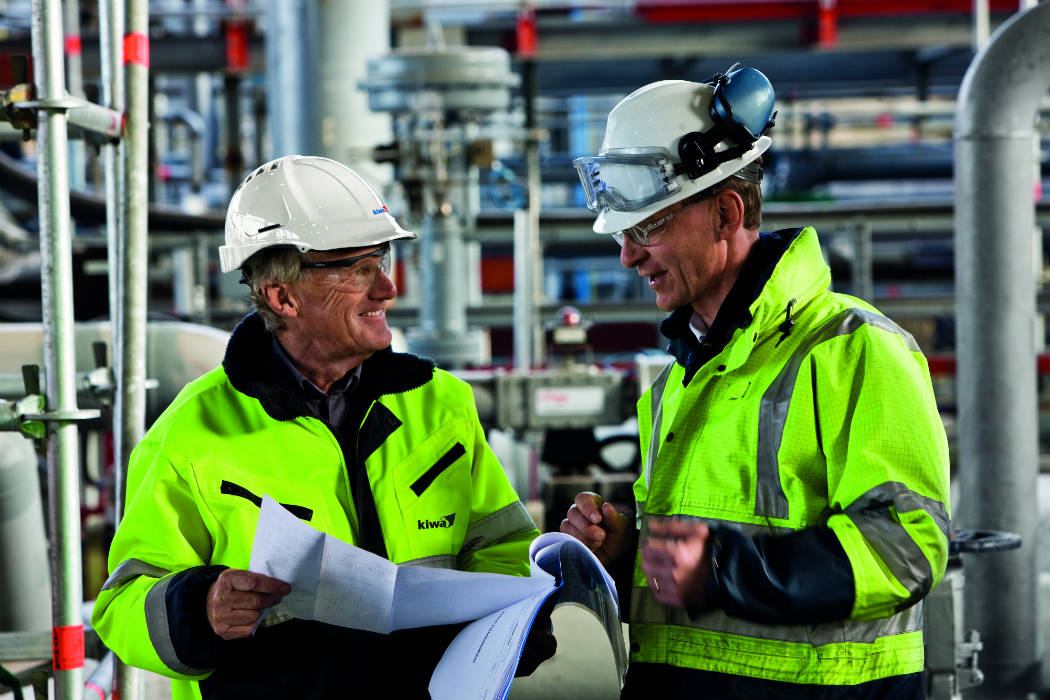 Two Kiwa engineers wearing bright yellow safety jackets and hard hats review blueprints in an industrial facility