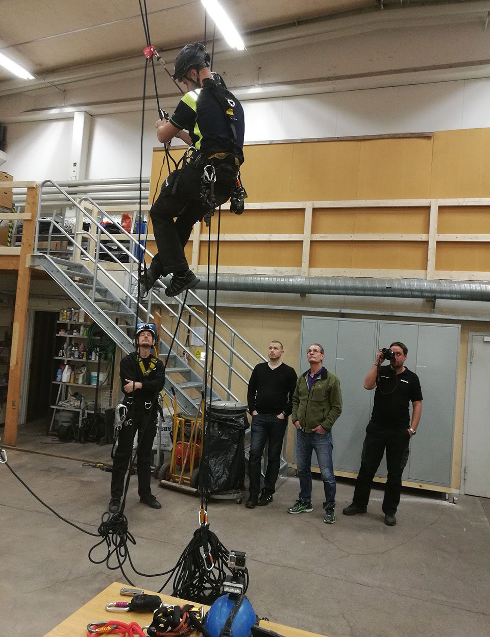 En person klättrar uppåt med utrustning för fallskydd i en industriell miljö, medan andra står och observerar.