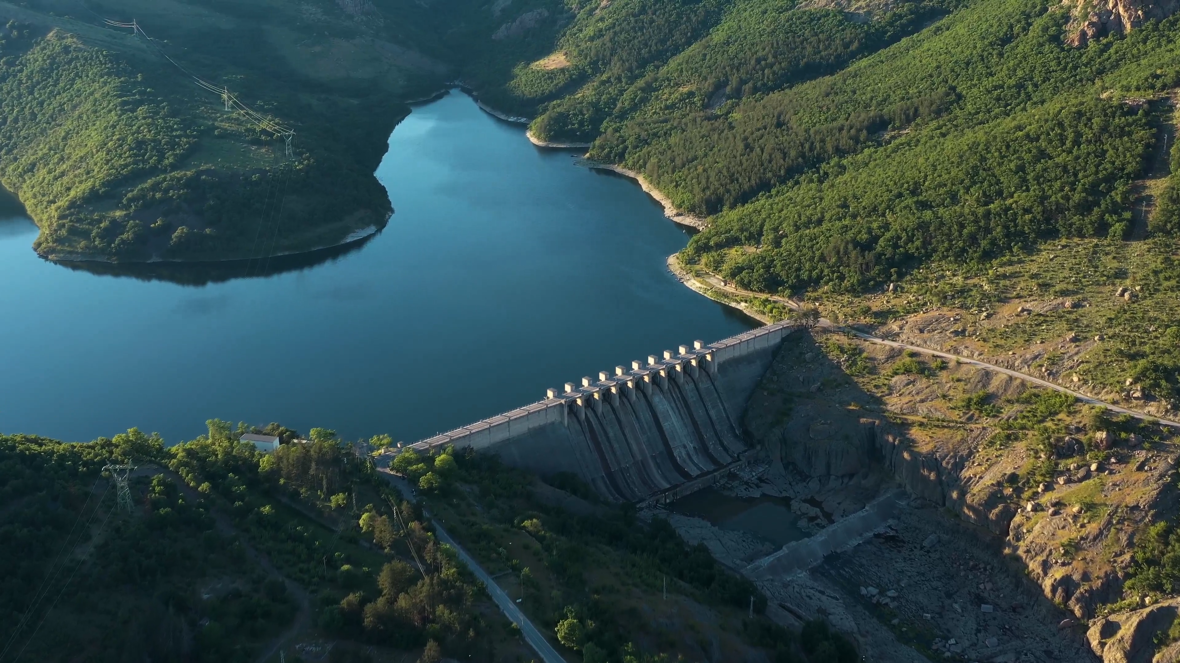 Aerial view of a dam surrounded by lush green hills and a serene river, showcasing a scenic landscape with a reservoir and forested areas