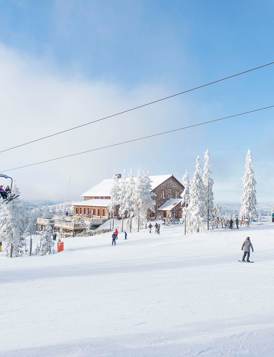 Snötäckt skidbacke med personer som åker skidor och snowboard, omgiven av frosttäckta träd och en träbyggnad i bakgrunden under en klar himmel.