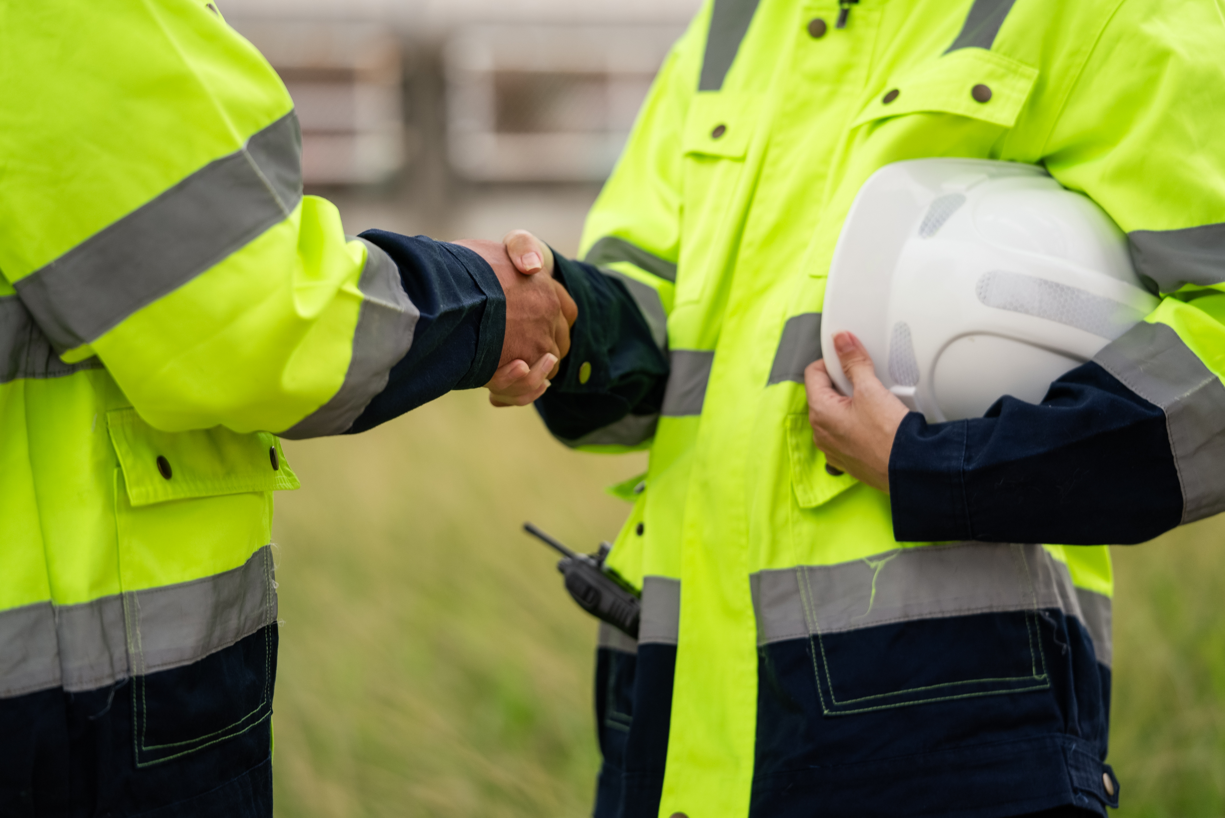 Two construction workers in high-visibility jackets shaking hands, with one holding a white hard hat.
