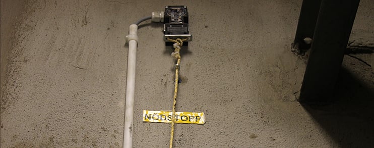 Nödstoppsknapp med gul varningsskylt på betonggolv, ansluten via kablar och rör.