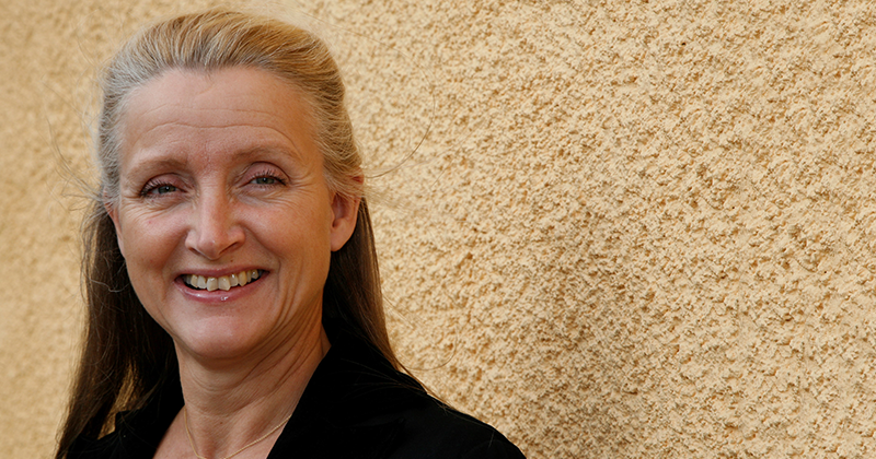 Kvinna med långt blont hår står framför en beige texturerad vägg. Hon har på sig en svart skjorta.