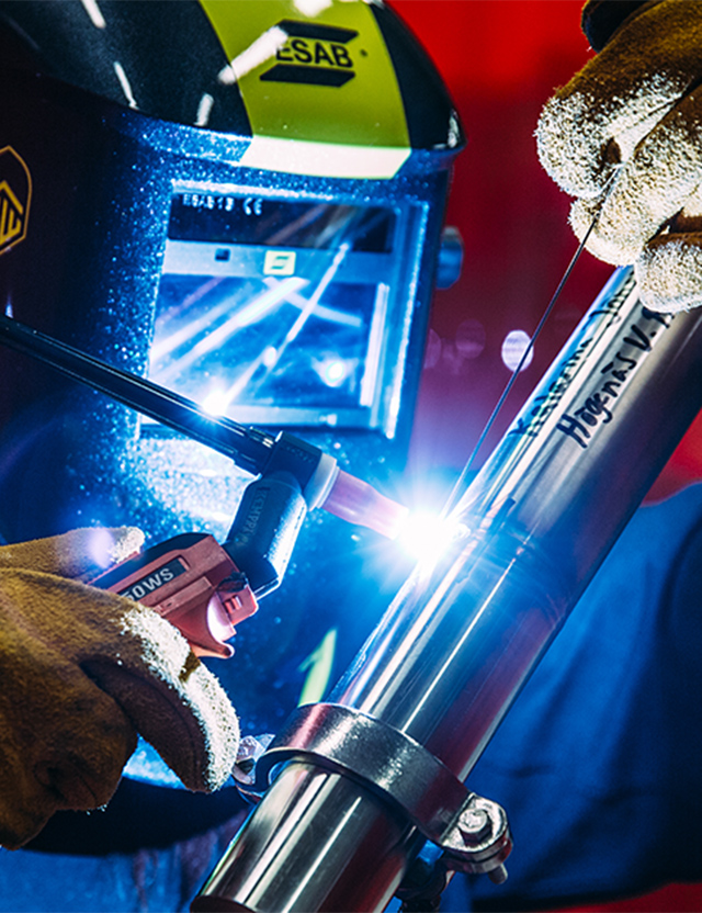Person som utför TIG-svetsning med skyddshjälm och handskar, svetsar en metallstång i starkt ljus.
