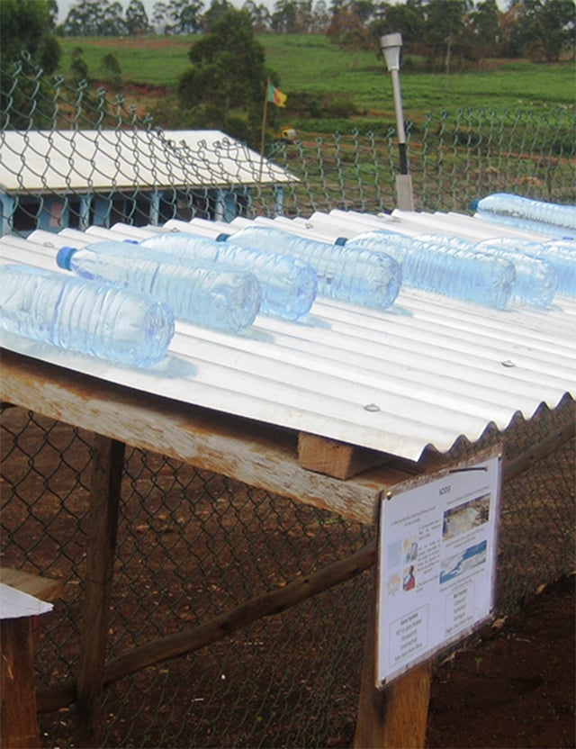 Plastflaskor fyllda med vatten placerade på ett korrugerat tak av metall för att värmas upp av solen, omgiven av ett stängsel och grönska.