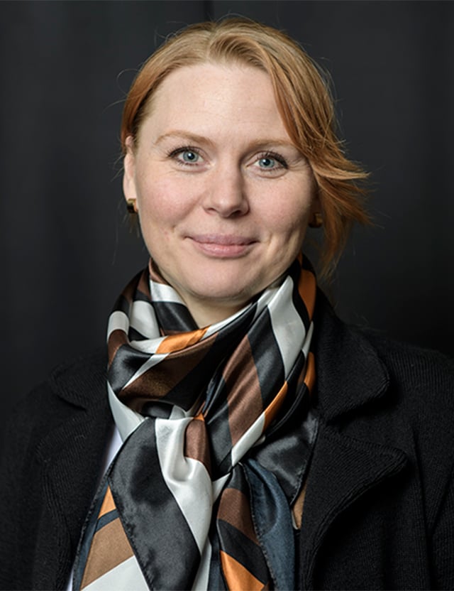 Person med ljust hår, klädd i en mörk kappa och en flerfärgad halsduk, framför en mörk bakgrund.