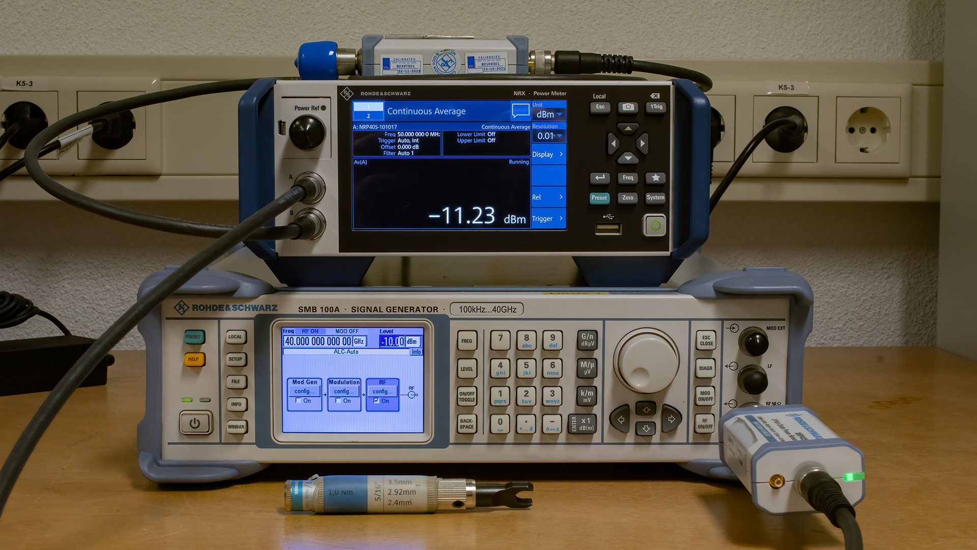Rohde & Schwarz SMB100A signal generator and NRP power sensor on a workbench, with digital screens displaying measurement readings, cables connected, and power outlets in the background. Ideal for electronics testing and measurement applications