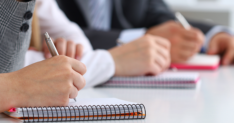 Personer som skriver i anteckningsböcker med pennor vid ett bord.