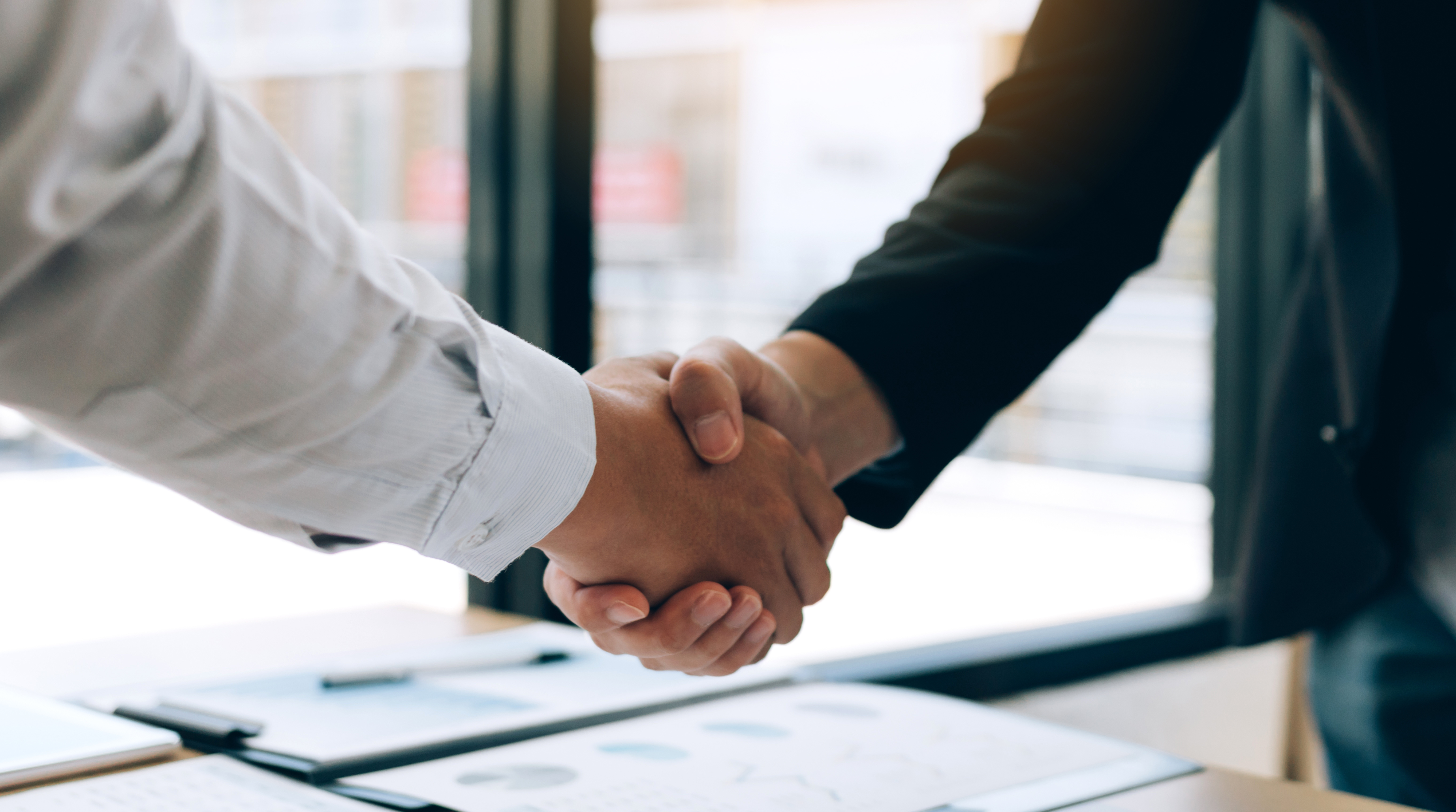 Two people shaking hands over a desk with documents, near a large window.