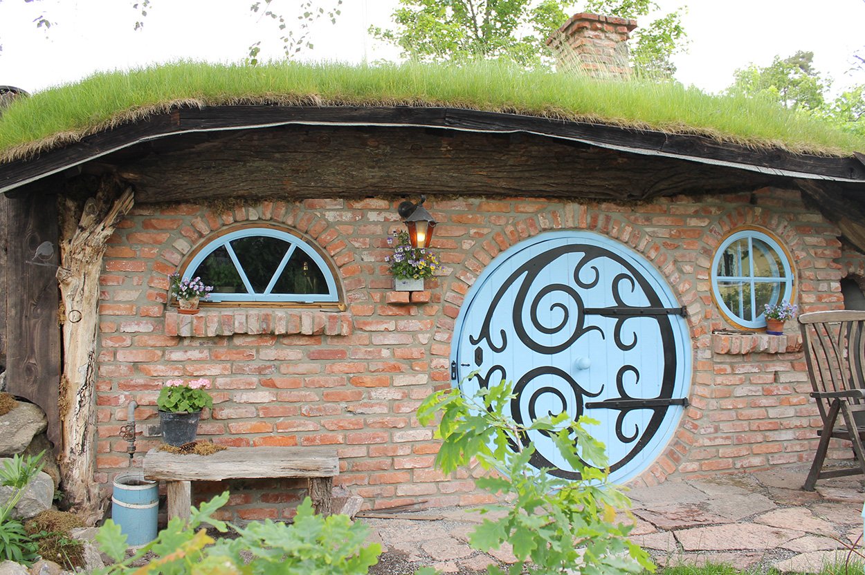 Tegelfasad med ett runt, blått dörr och grästak, omgiven av växtlighet.