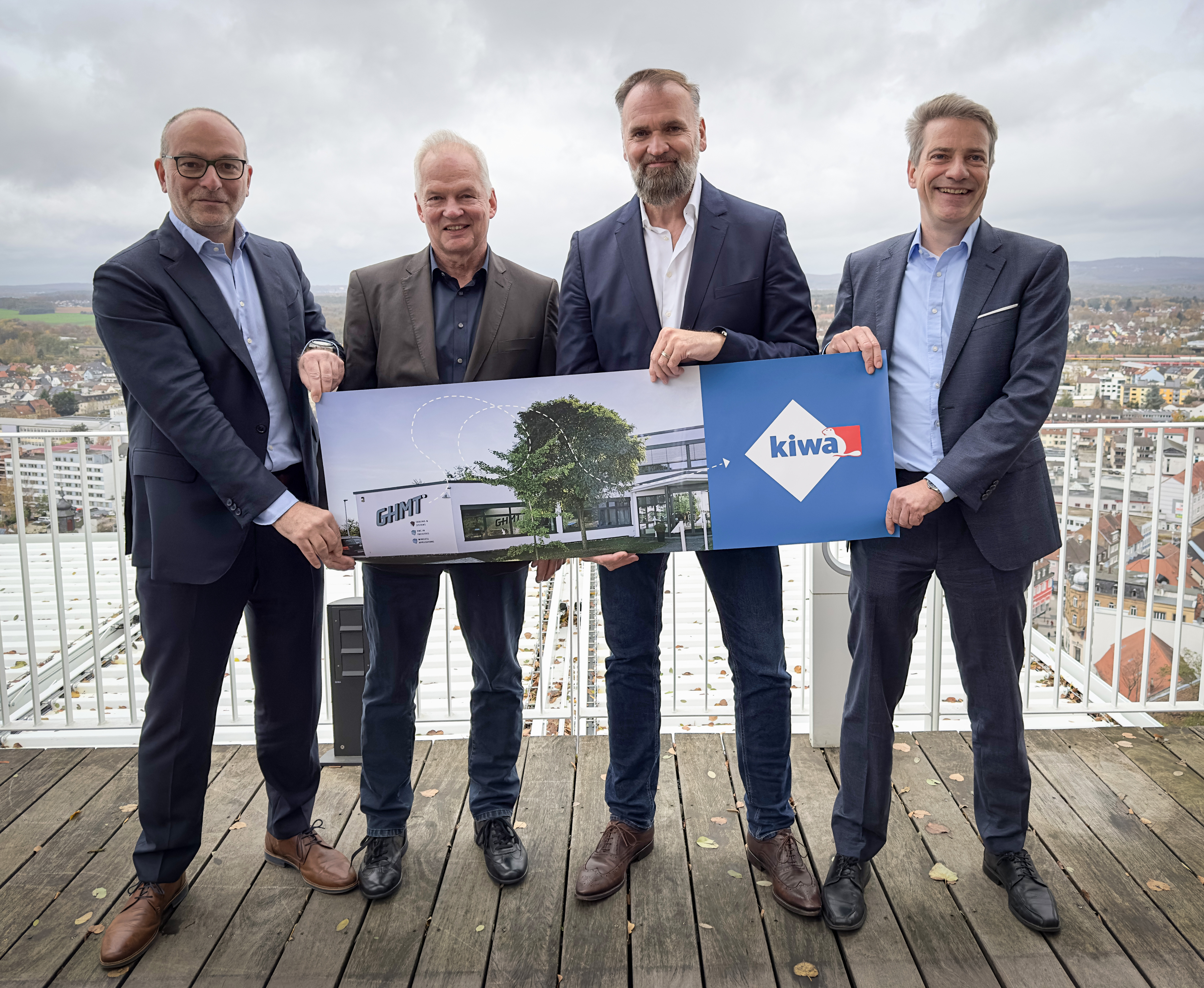 Four senior managers in business attire holding a poster with a building image and a Kiwa logo, standing on a balcony with a cityscape in the background
