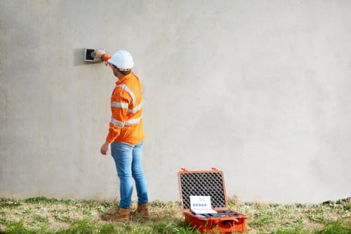 Kiwa inspector using a handheld device to inspect a concrete wall, wearing an orange safety jacket and white hard hat, with an open equipment case nearby