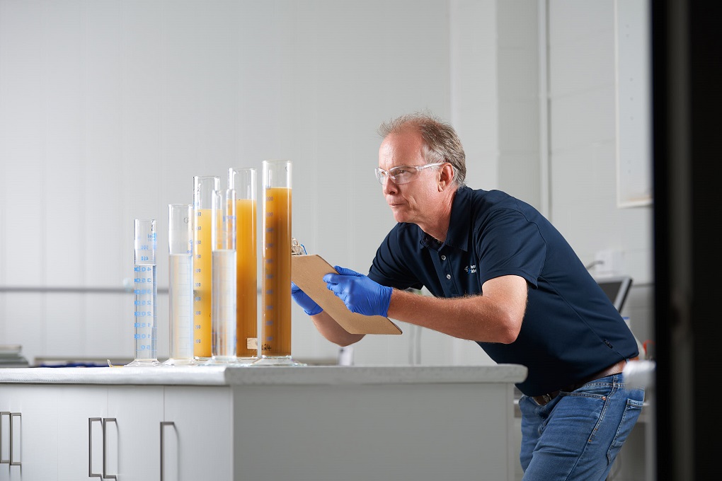 Kiwa laboratory employee examining test tubes with liquid samples in a laboratory, wearing safety goggles and gloves, holding a clipboard for notes