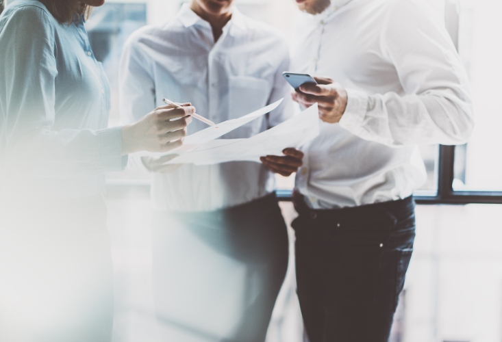 Three business professionals collaborating, examining documents and using a smartphone in a brightly lit office setting