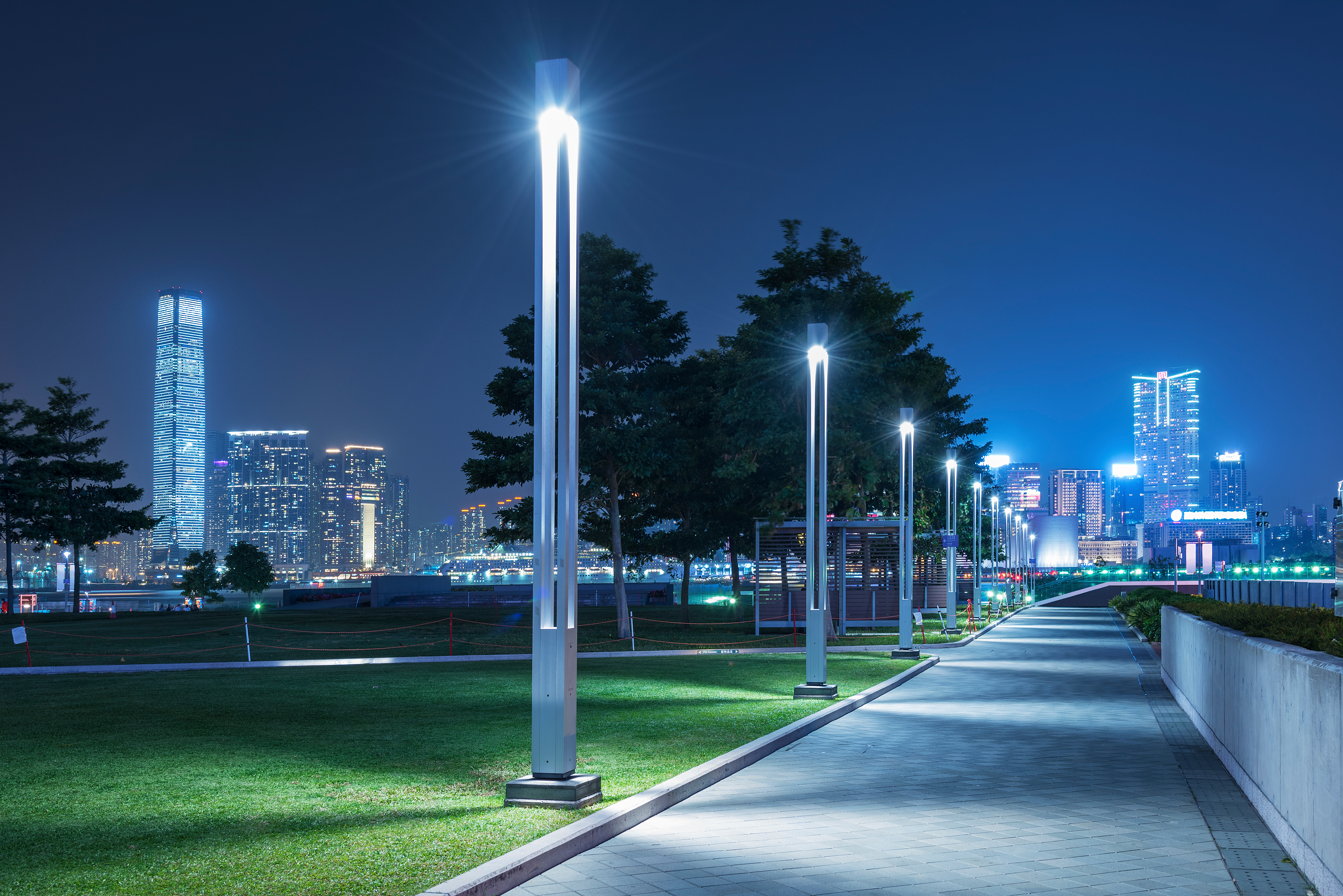 Night view of a cityscape with illuminated skyscrapers and a well-lit park pathway, featuring modern street lamps and green space in the foreground