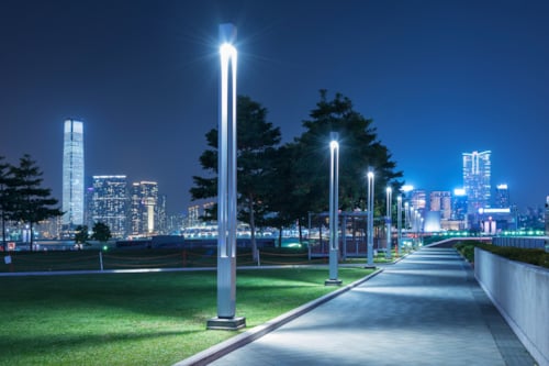 Night view of a cityscape with illuminated skyscrapers and a well-lit park pathway, featuring modern street lamps and green space in the foreground