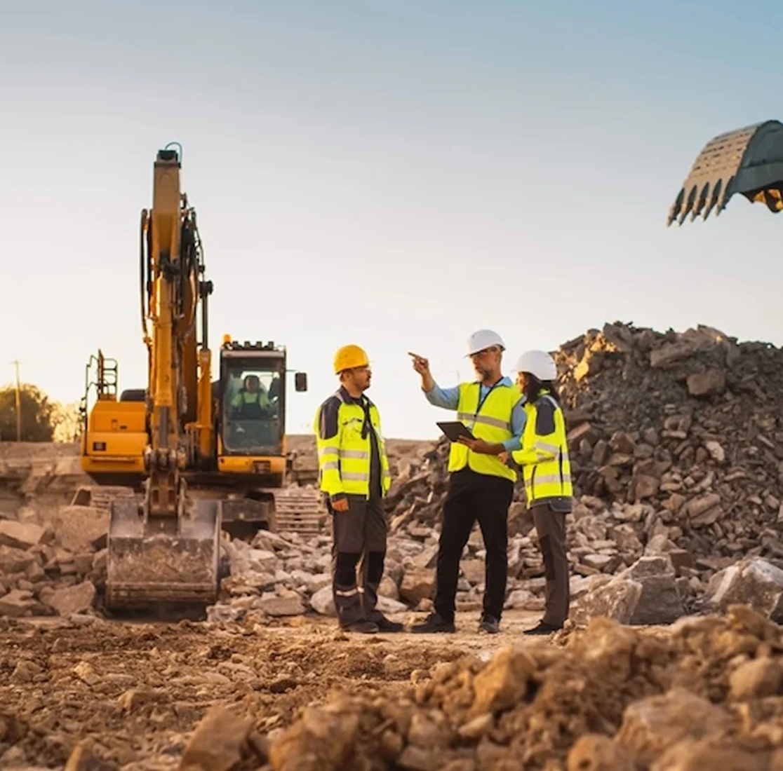 Construction site with three Kiwa auditors wearing safety vests and helmets discussing plan, standing next to a large excavator and rubble pile, under clear sky