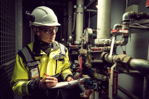 Kiwa engineer in safety gear inspecting industrial pipes and gauges, taking notes on a clipboard in a mechanical room