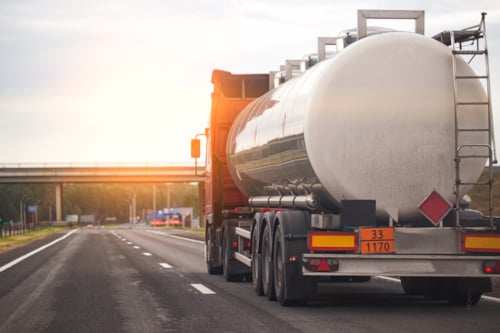 Tanker truck transporting liquid cargo on a highway during sunset, with a bridge in the background