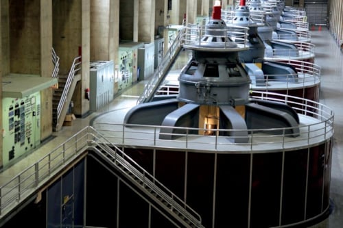 Rader av stora turbinmaskiner i en industriell anläggning med betongväggar, paneler och gångvägar.