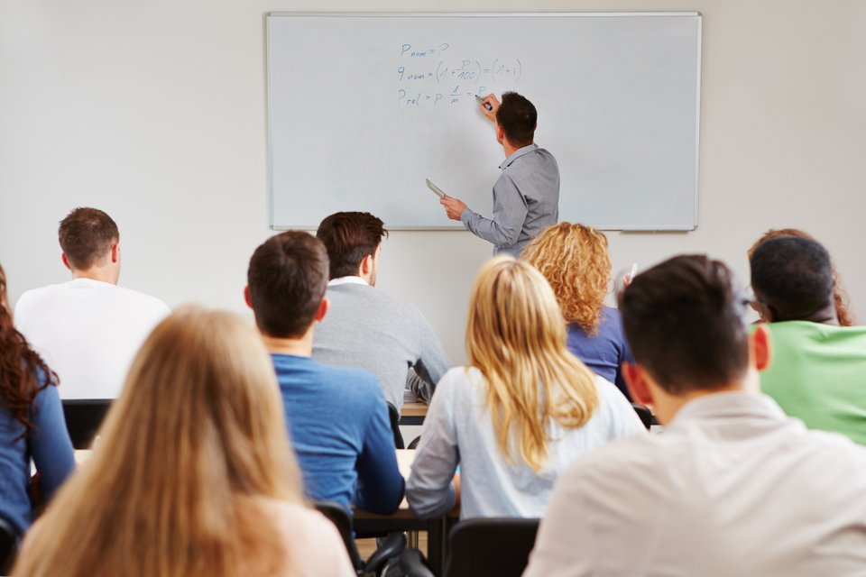 Teacher writing mathematical equations on a whiteboard in a classroom with attentive students seated, focused on learning