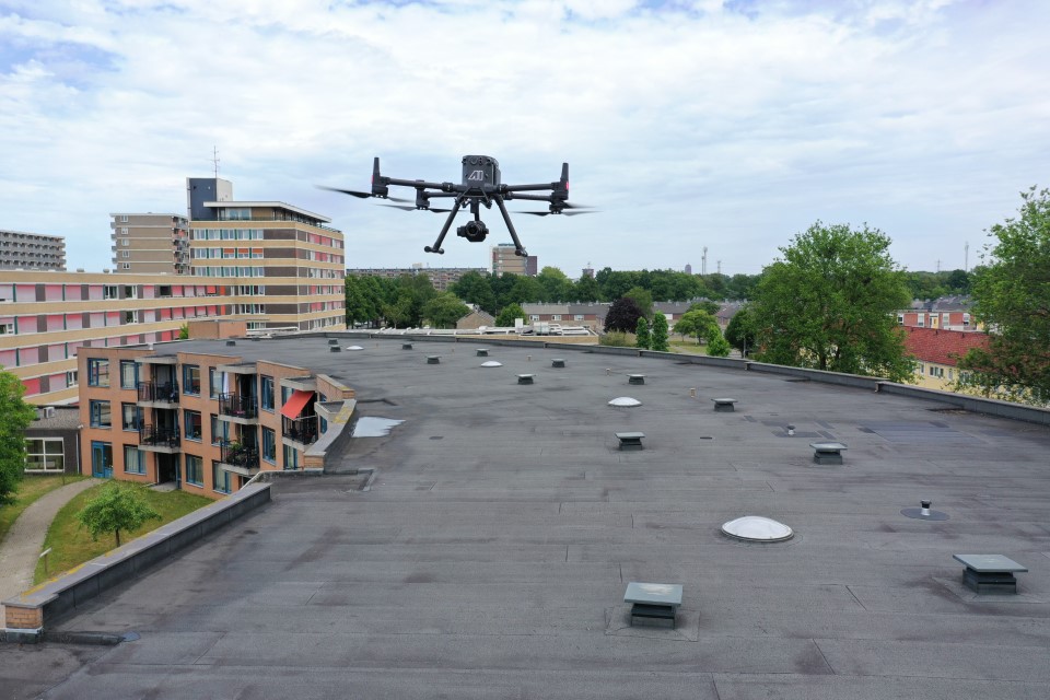 Drone vliegt boven een plat dak van een gebouw in een stedelijke omgeving met bomen en appartementen op de achtergrond.
