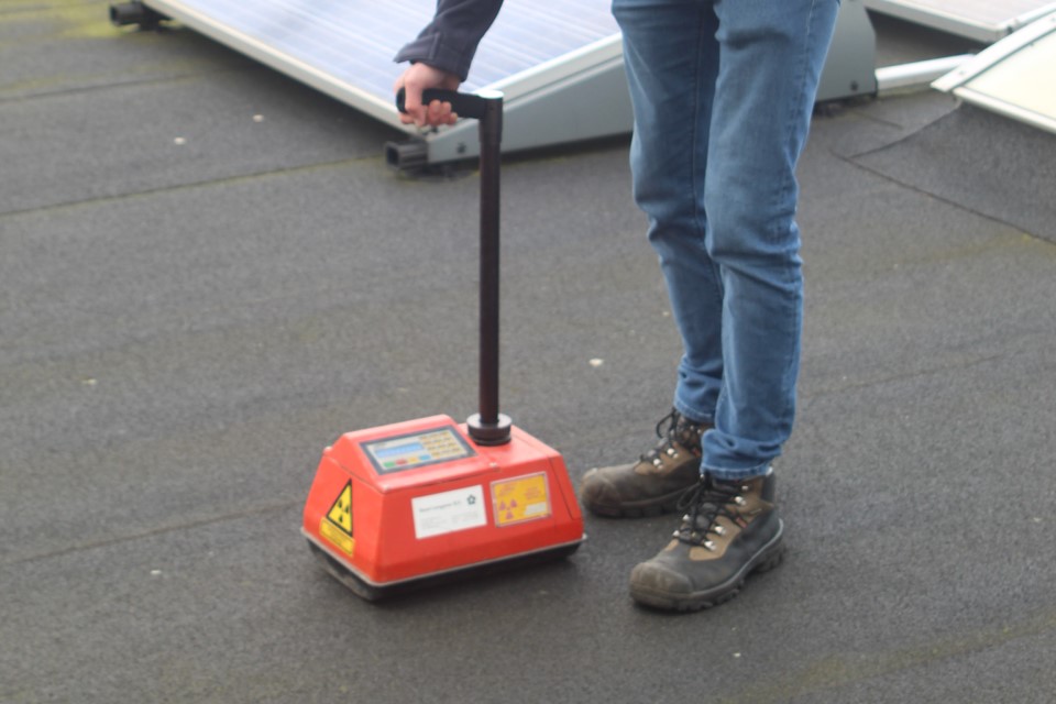 Persoon gebruikt een draagbaar apparaat met een rode behuizing en waarschuwingssymbolen op een donkere vloer, mogelijk op een plaats met zonnepanelen.