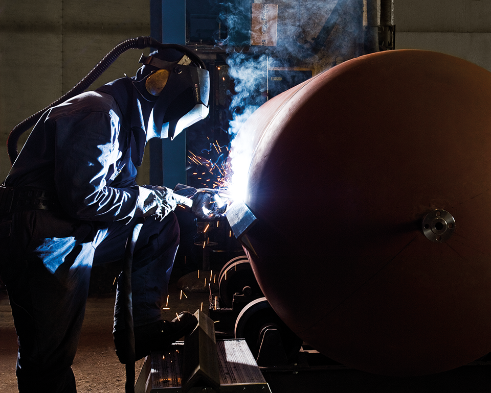 A Kiwa certified welder in protective gear working on large metal cylinder, emitting bright sparks, in industrial workshop