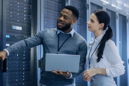 Two IT professionals in a data center discussing server maintenance while holding a laptop, surrounded by server racks