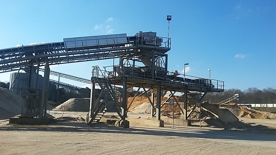 Een industriële zand- en grindverwerkingsinstallatie met transportbanden, metalen structuren en stapels materiaal onder een blauwe lucht.