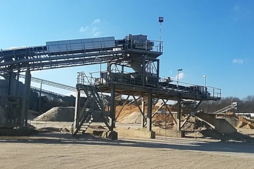 Een industriële zand- en grindverwerkingsinstallatie met transportbanden, metalen structuren en stapels materiaal onder een blauwe lucht.