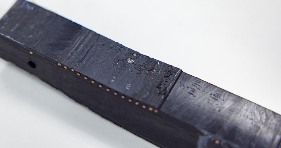 Zwart rechthoekig object met een matte afwerking, zichtbaar slijtage en kleine details op het oppervlak, op een witte achtergrond.