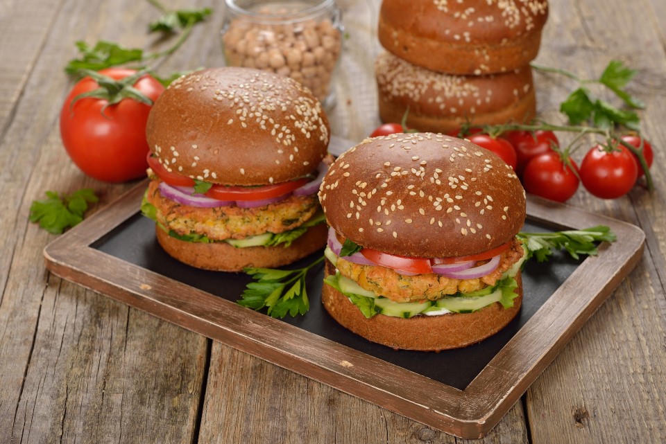 Twee vegetarische hamburgers op een houten plank, gegarneerd met tomaat, rode ui, komkommer en peterselie, omringd door verse groenten en broodjes.