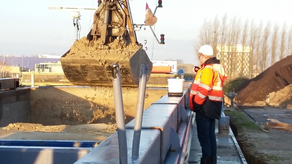 Man in een oranje veiligheidsjas en witte helm observeert een graafmachine die zand verplaatst op een bouwplaats.