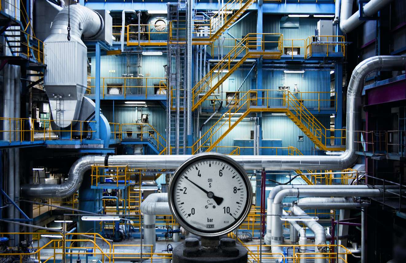Tryckmätare i förgrunden av en industriell anläggning med rör och metallstrukturer.