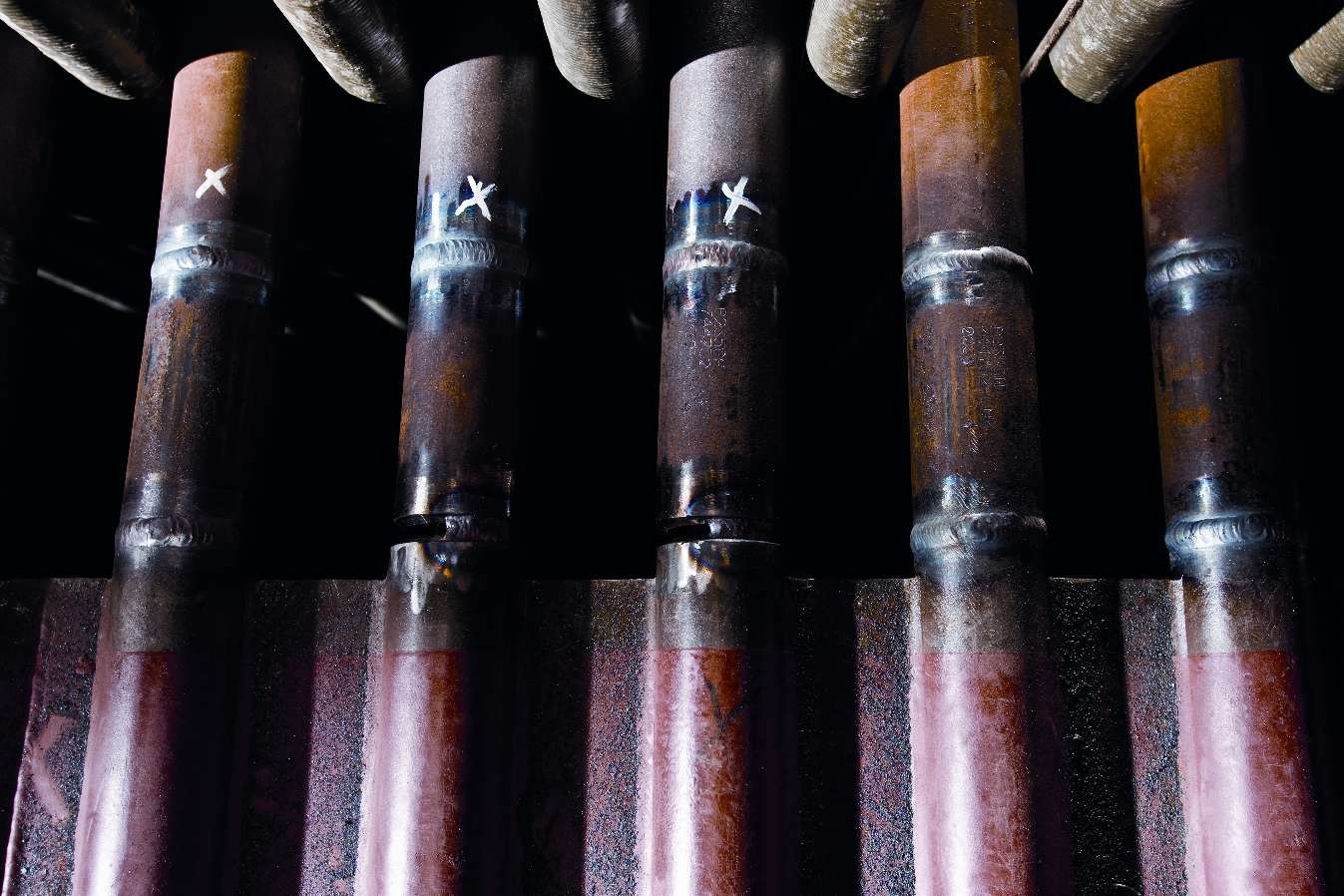 Närbild av flera rostiga metallrör med svetsskarvar och märkning, arrangerade vertikalt i industriell miljö.