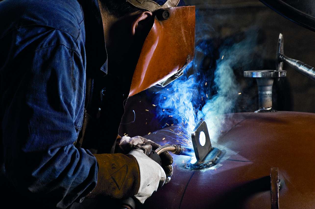 Svetsare i blå arbetskläder och skyddsutrustning som svetsar metall, med gnistor och ett blått sken synligt.