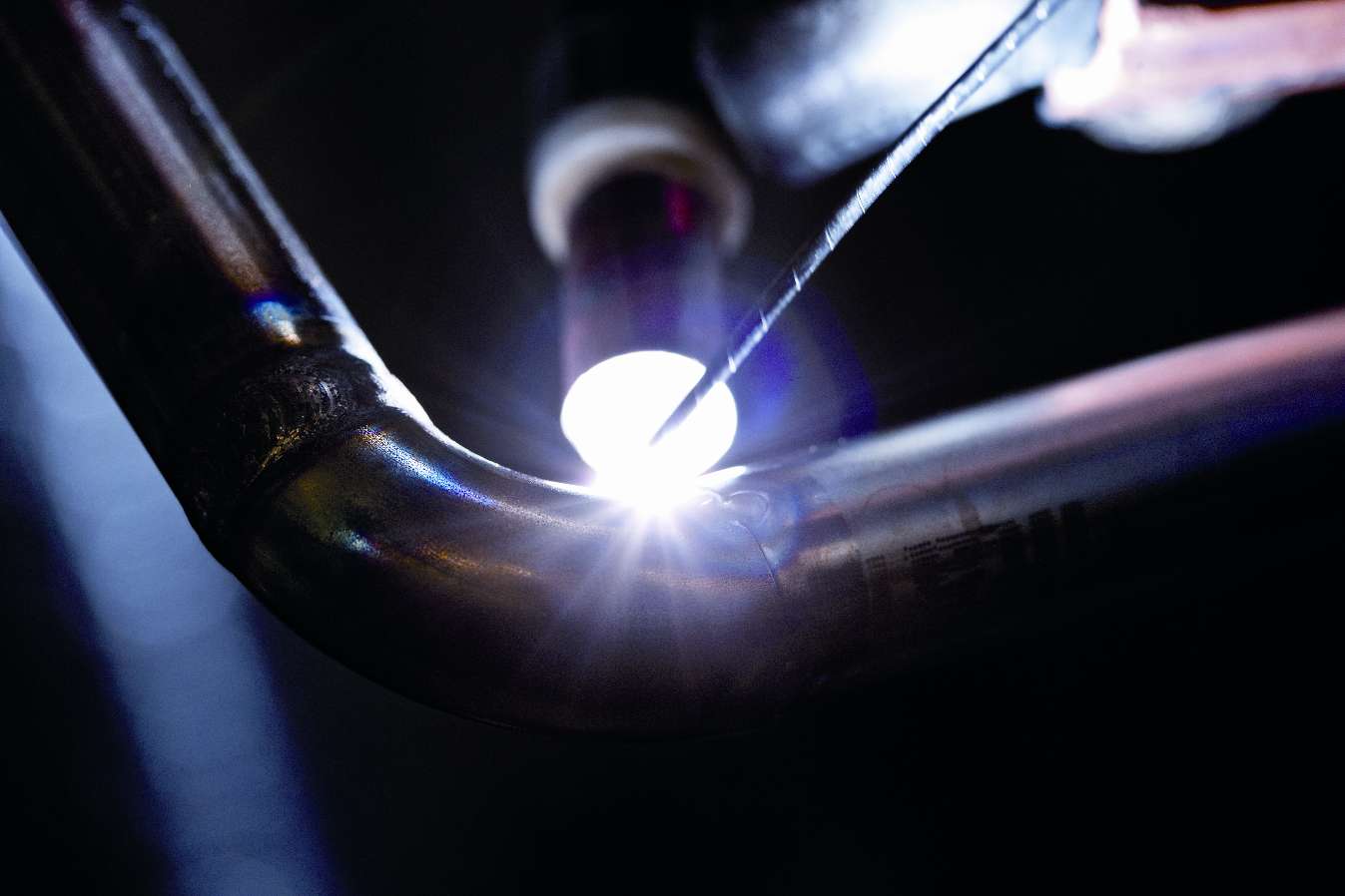 Närbild av en svetssöm på ett metallrör under TIG-svetsning, med ljusbågen i fokus och gnistor synliga.