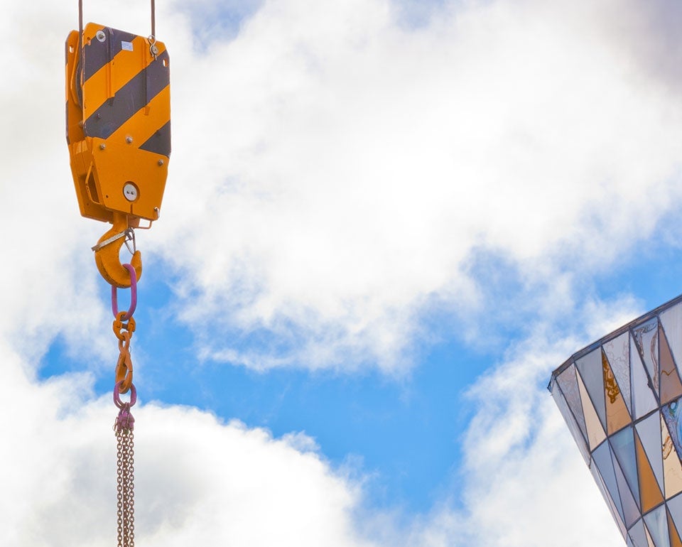 Lyftkran med gul/svart randig talja med kedja för tunga lyft och säker lasthantering, hänger mot en bakgrund av molnig himmel, med en del av en glaskupol i hörnet.