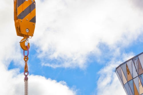 Lyftkran med gul/svart randig talja med kedja för tunga lyft och säker lasthantering, hänger mot en bakgrund av molnig himmel, med en del av en glaskupol i hörnet.