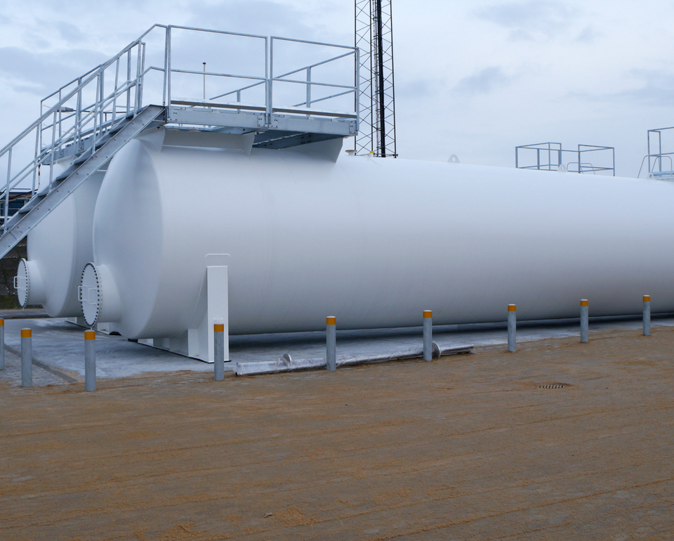 Vit cylindrisk tank utomhus på en industriell plats, omgiven av stolpar och med en trappa som leder upp till toppen.