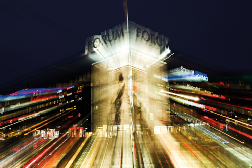 Byggnad med texten "Forum" och "Första" på fasaden, upplyst mot en mörk kvällshimmel, omgiven av ljusspår från rörelse och neonljus.