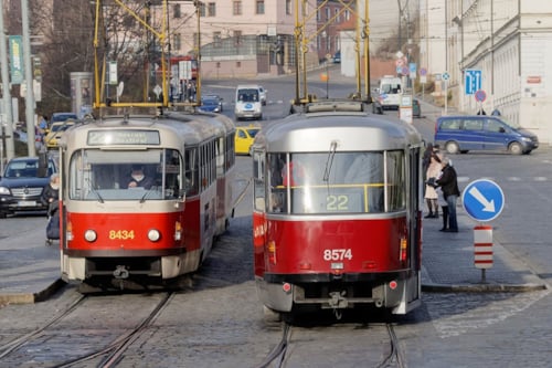 Två röda och grå spårvagnar på en stadsgata med korsande spår och omgivande byggnader i bakgrunden.
