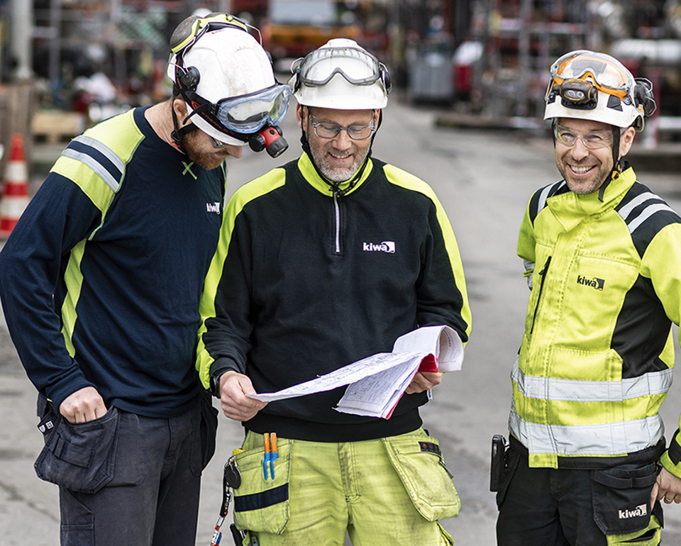 Tre besiktningsingenjörer i skyddsutrustning som diskuterar en ritning på en industrianläggning.