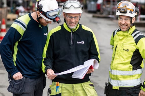 Tre besiktningsingenjörer i skyddsutrustning som diskuterar en ritning på en industrianläggning.