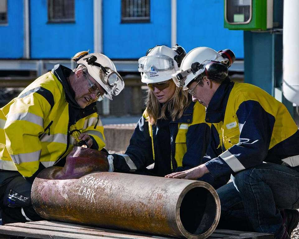 En erfaren inspektör lyser med ficklampa in i ett stålrör, två fokuserade personer står bredvid för att ta del av informationen. Samtliga har hjälm och skyddskläder på sig. 