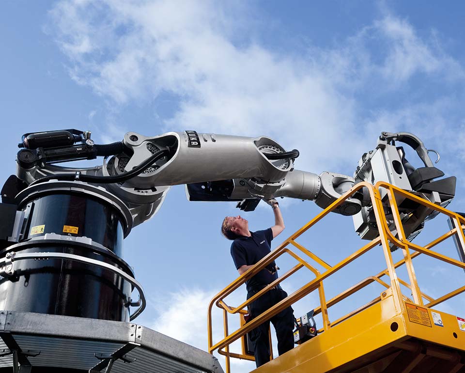 En man arbetar med en stor industrirobotarm utomhus, stående på en gul plattform under en klar himmel.