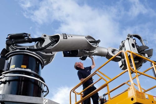 En man arbetar med en stor industrirobotarm utomhus, stående på en gul plattform under en klar himmel.