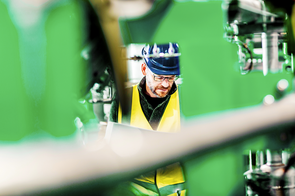 En person i gul reflexväst och blå hjälm arbetar bland industrimaskiner.