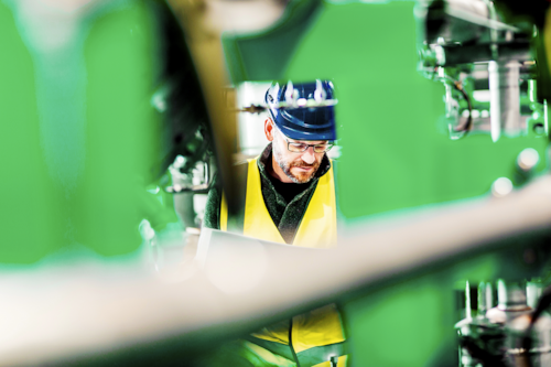En person i gul reflexväst och blå hjälm arbetar bland industrimaskiner.